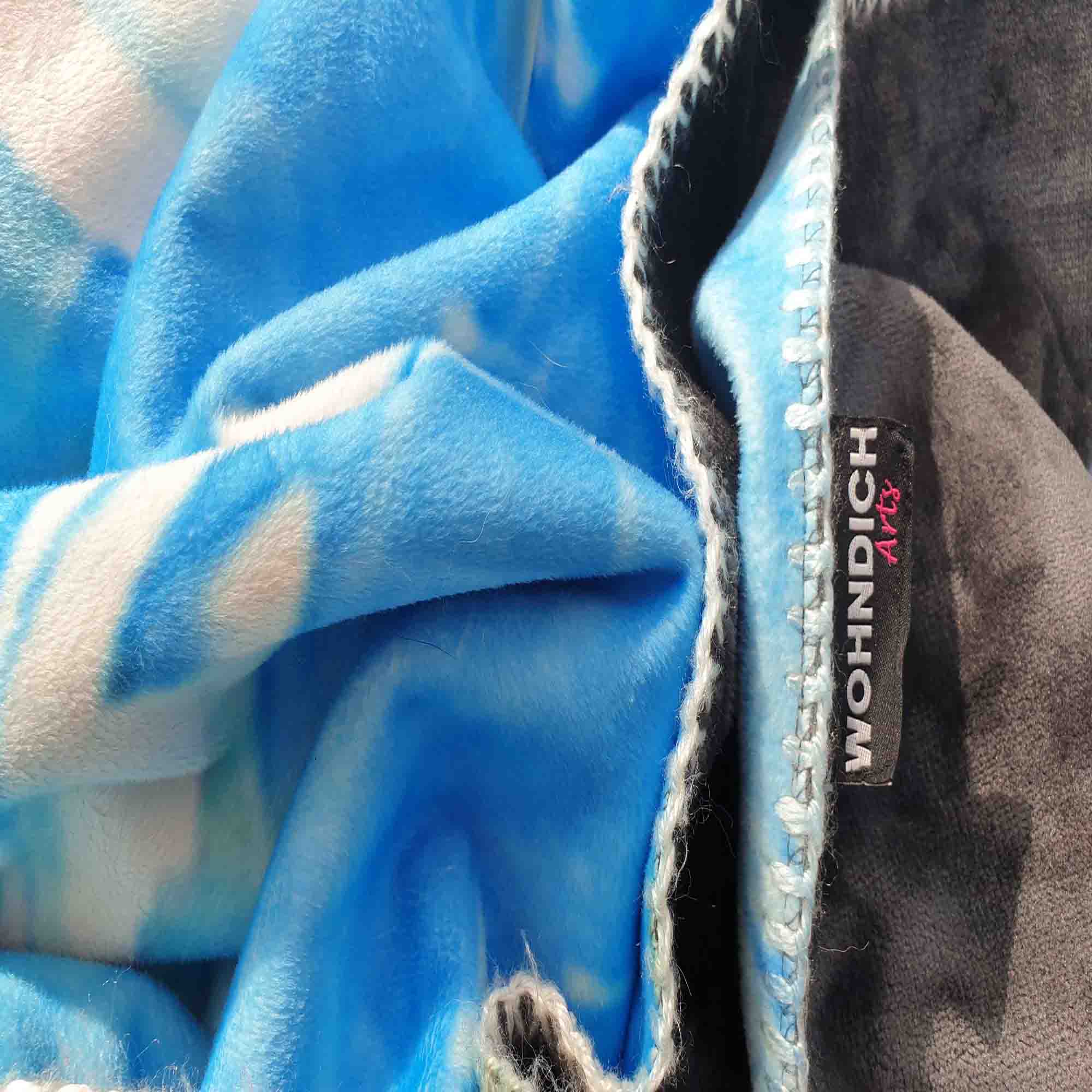 Detailaufnahme einer blauen Kuscheldecke mit weißer Kettelnaht und WOHNDICH Arts-Label.
