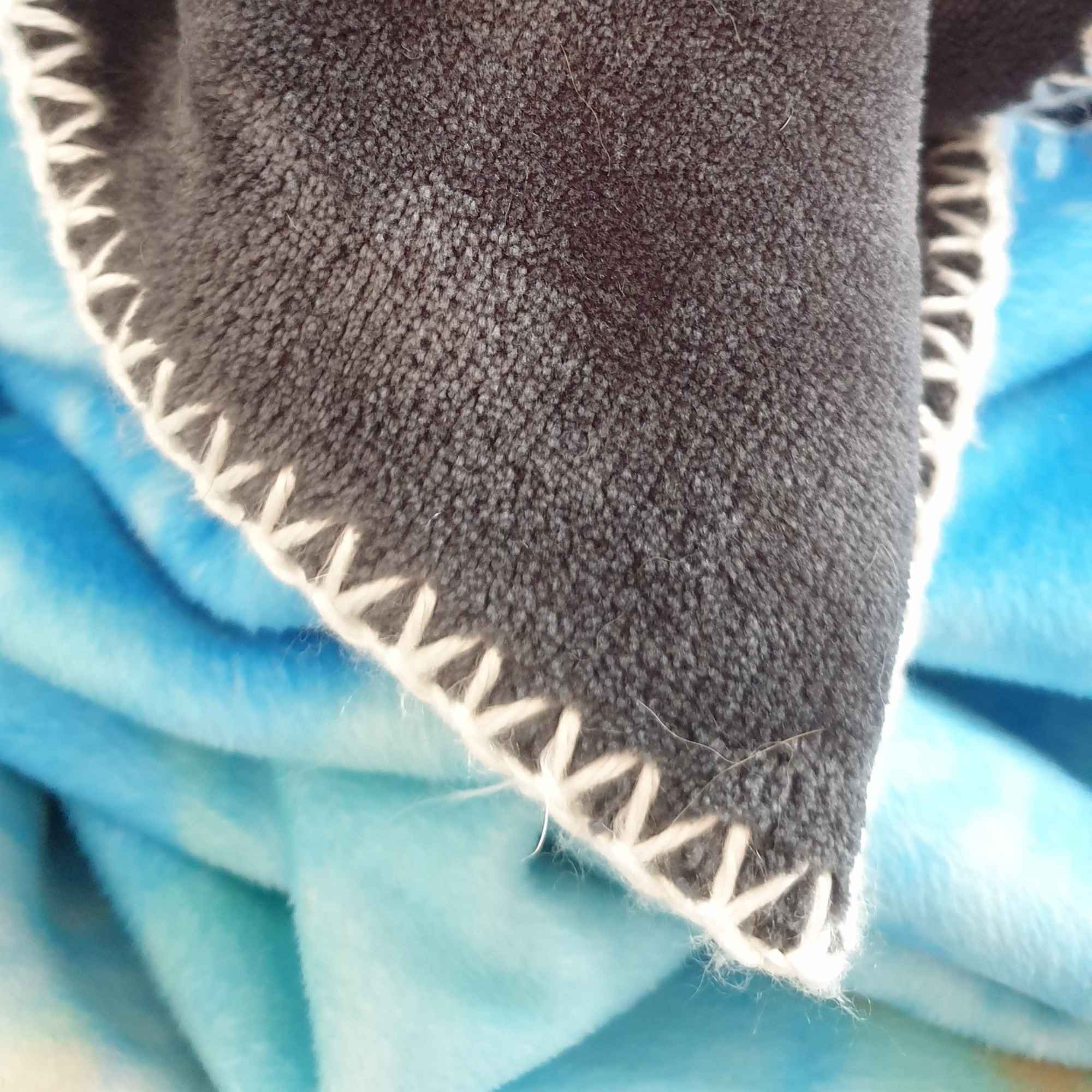 Detailaufnahme der flauschigen Kuscheldecke mit schwarzer Rückseite, heller Ziernaht und himmelblauem Untergrund.