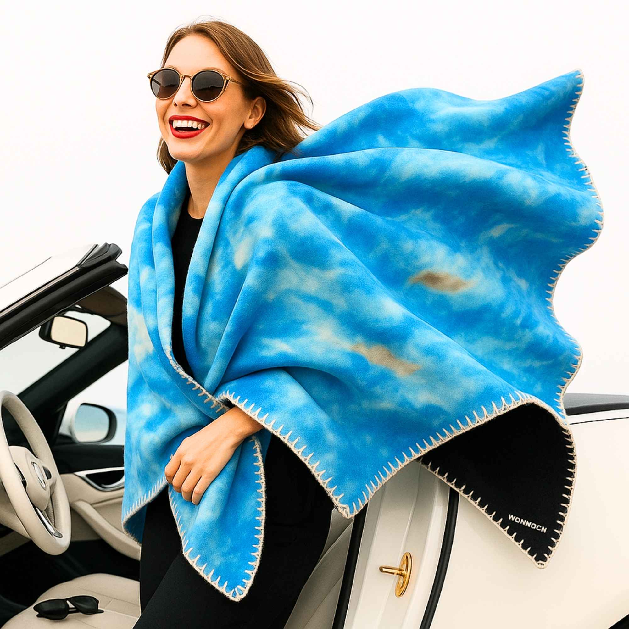 Frau mit Sonnenbrille lacht und trägt eine himmelblaue Kuscheldecke mit heller Kettelnaht über den Schultern, lehnt an einem Cabrio.