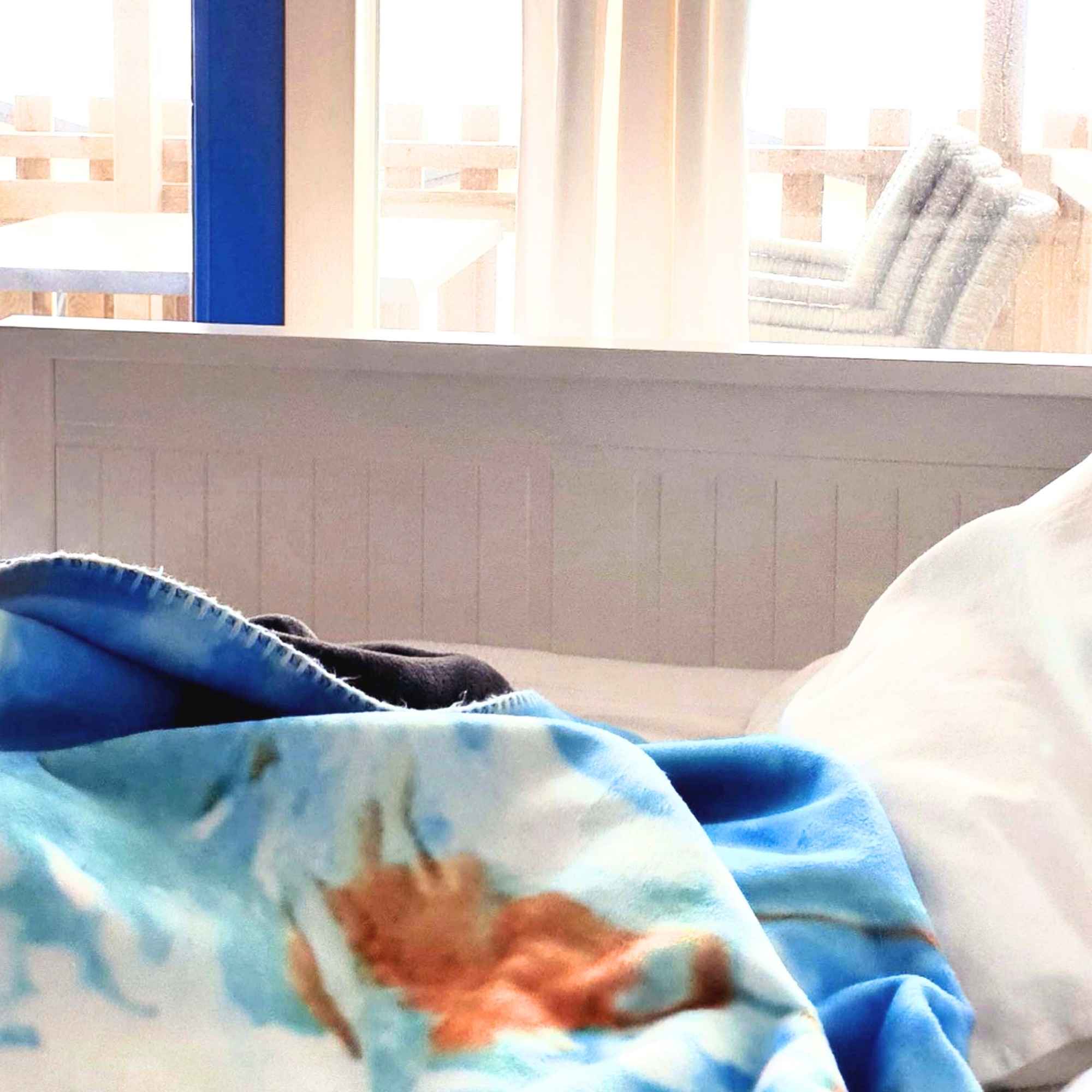 Himmelblaue Kuscheldecke mit weiß-blauen und ockerfarbenen Wolkenmustern liegt auf einem Bett vor einem hellen Fenster.