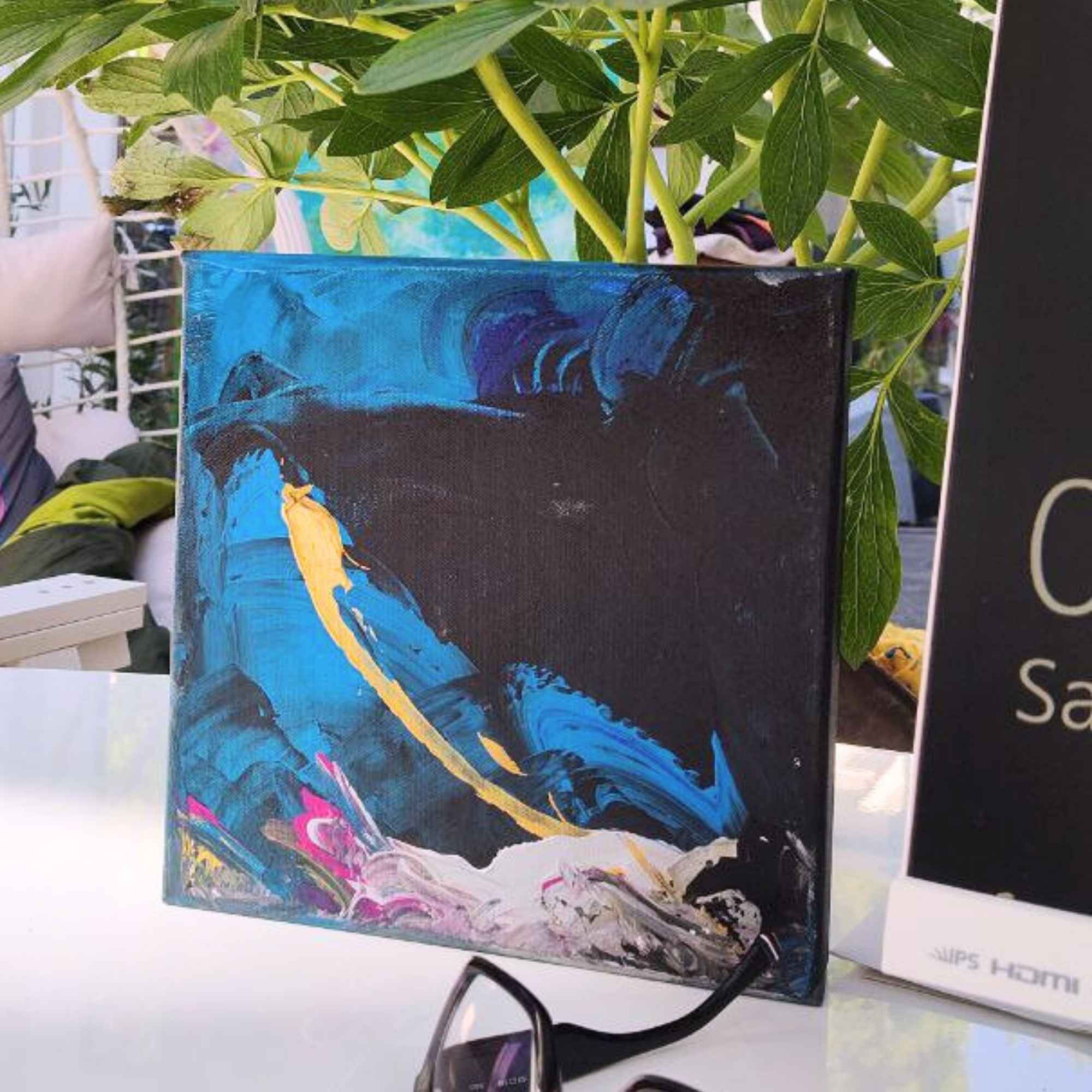 Ein kleines, abstraktes Acrylbild in Blau, Schwarz und Gold steht auf einem weißen Tisch. Im Hintergrund eine grüne Pflanze, daneben eine Sonnenbrille und ein Schild mit Text. Das Bild zeigt eine dynamische goldene Linie auf dunklem Grund – wie eine Sternschnuppe. Künstlerin: Regine Rauin