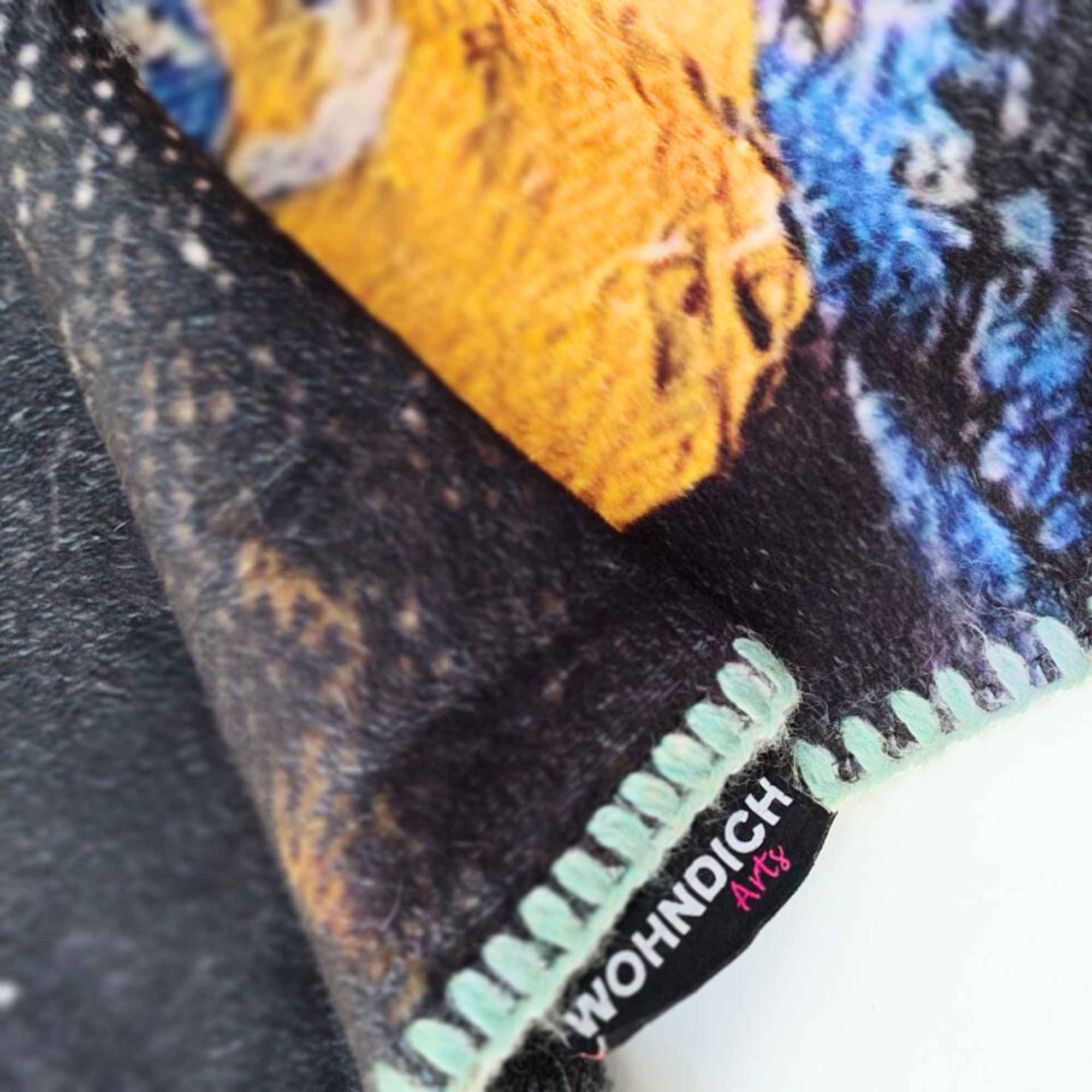 Detailaufnahme einer Kuscheldecke mit sichtbarem „WOHNDICH Arts“-Label, farbigem Kettelrand und strukturreichem Druck in Ocker, Blau und Schwarz.