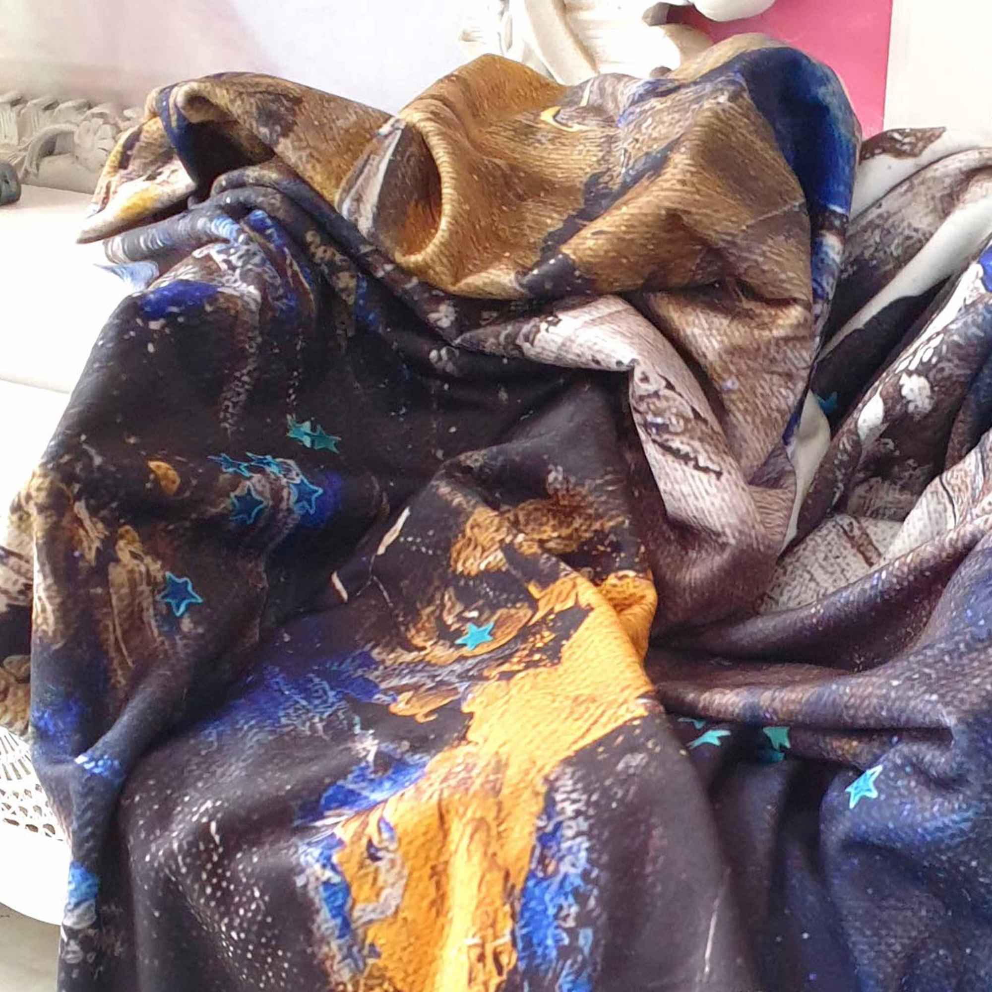 Kuscheldecke mit abstraktem Druck in Dunkelblau, Braun, Ocker und tĂĽrkisfarbenen Sternen, locker drapiert auf einem weiĂźen Stuhl.