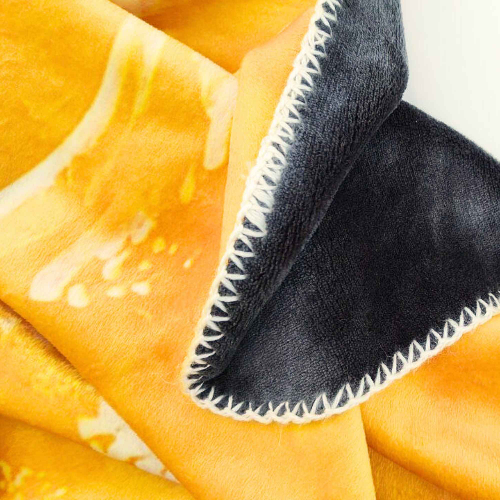 Detailansicht der Kuscheldecke HEALING mit orange-gelbem Farbverlauf und dunkelgrauer Rückseite, weißer Zickzack-Naht als sichtbares Designelement.
