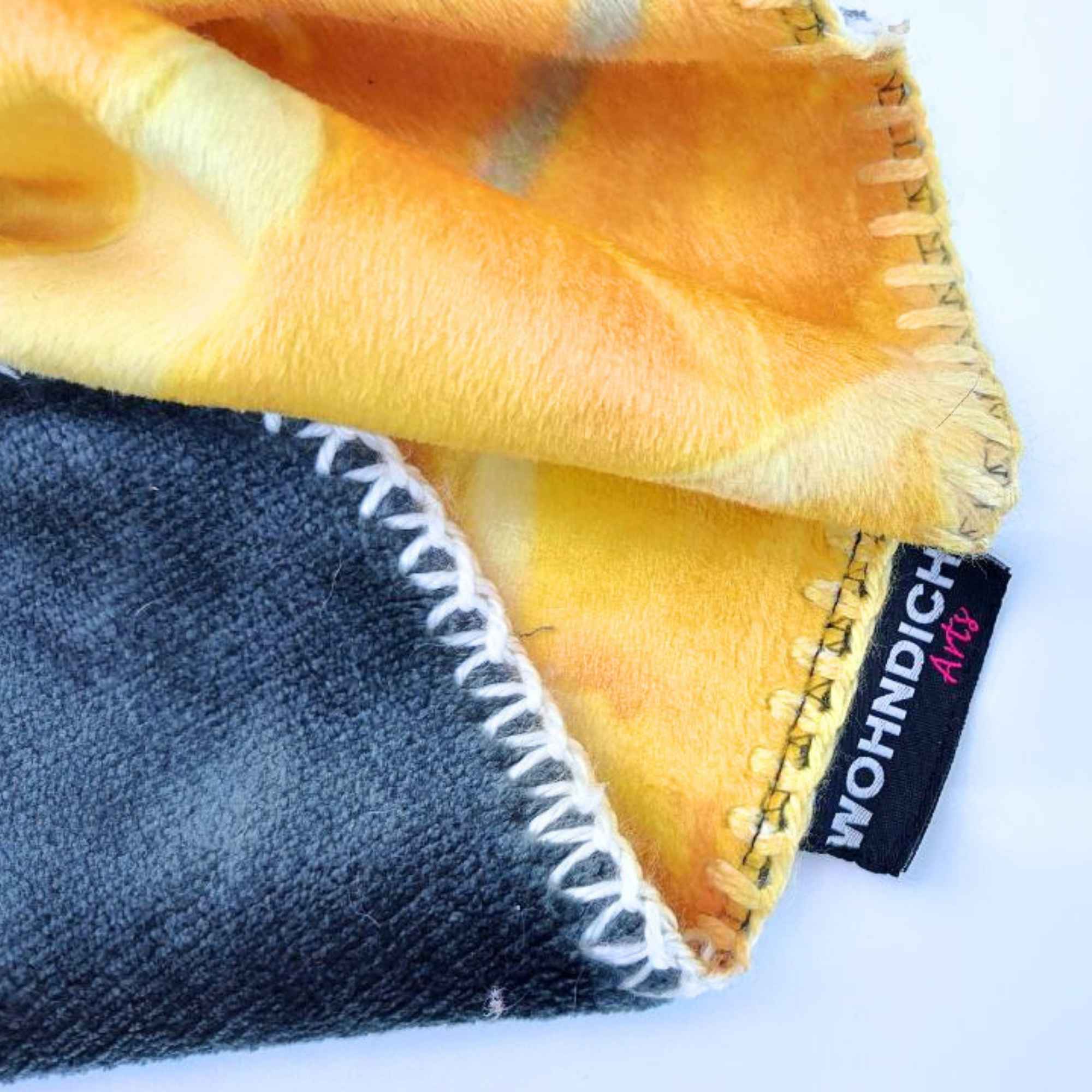 Kuscheldecke mit kontrastreicher Rückseite und dekorativer Naht; das Markenlabel „WOHNDICH ART“ ist sichtbar am unteren Rand vernäht.