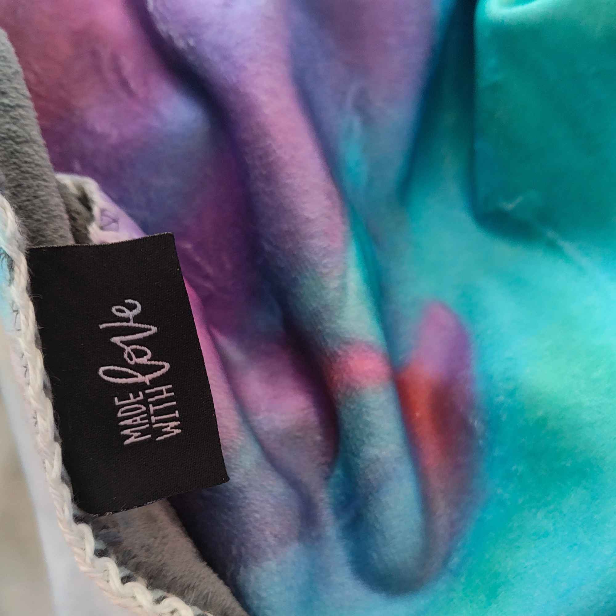Detail der weichen Kuscheldecke FORREST GUMP mit „Made with LOVE“-Label und abstraktem Motiv in Türkis, Blau, Violett und Rot.