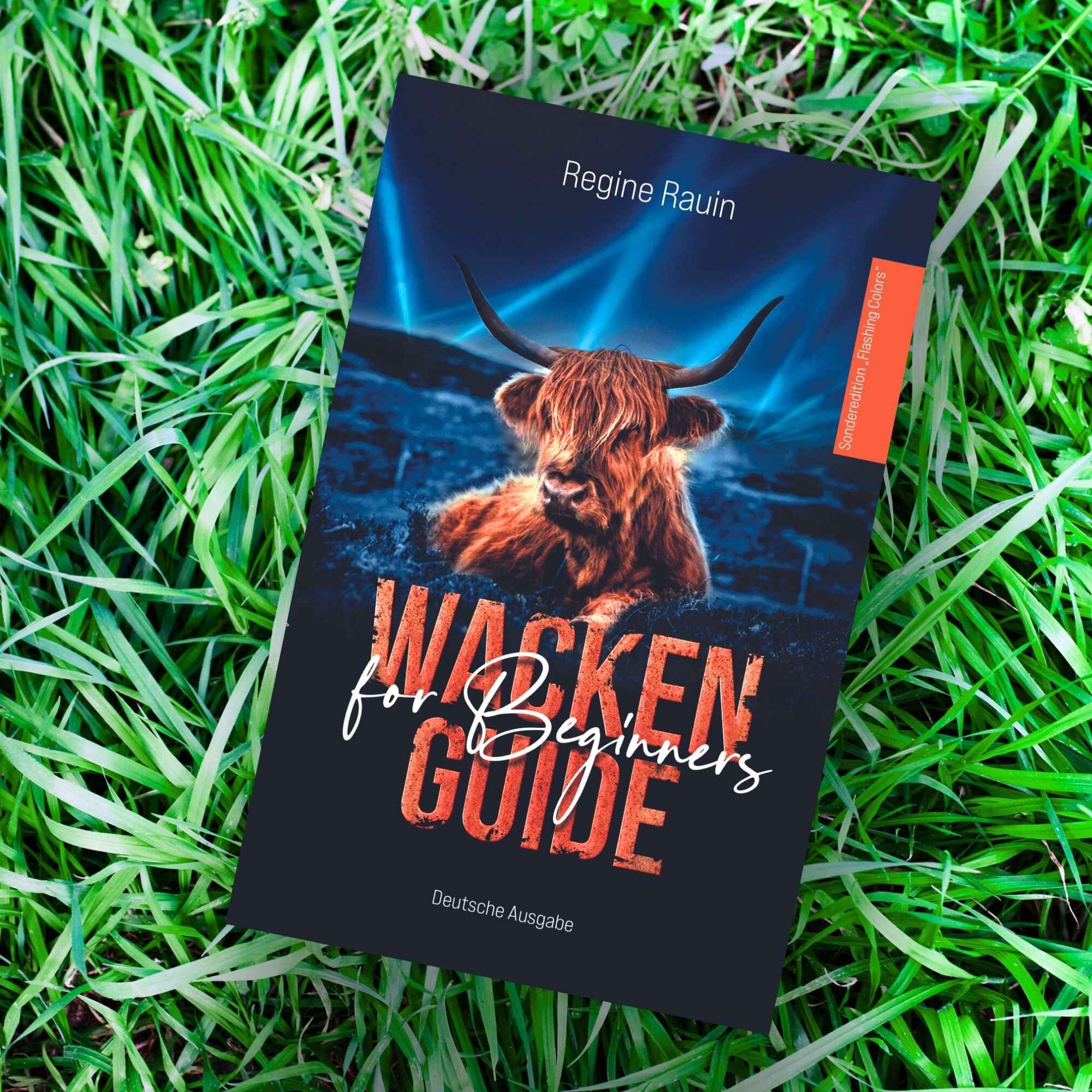 „Buch mit Titel ‚Wacken Guide for Beginners‘ auf Gras, Cover mit Hochlandrind und Bühnenscheinwerfern.“