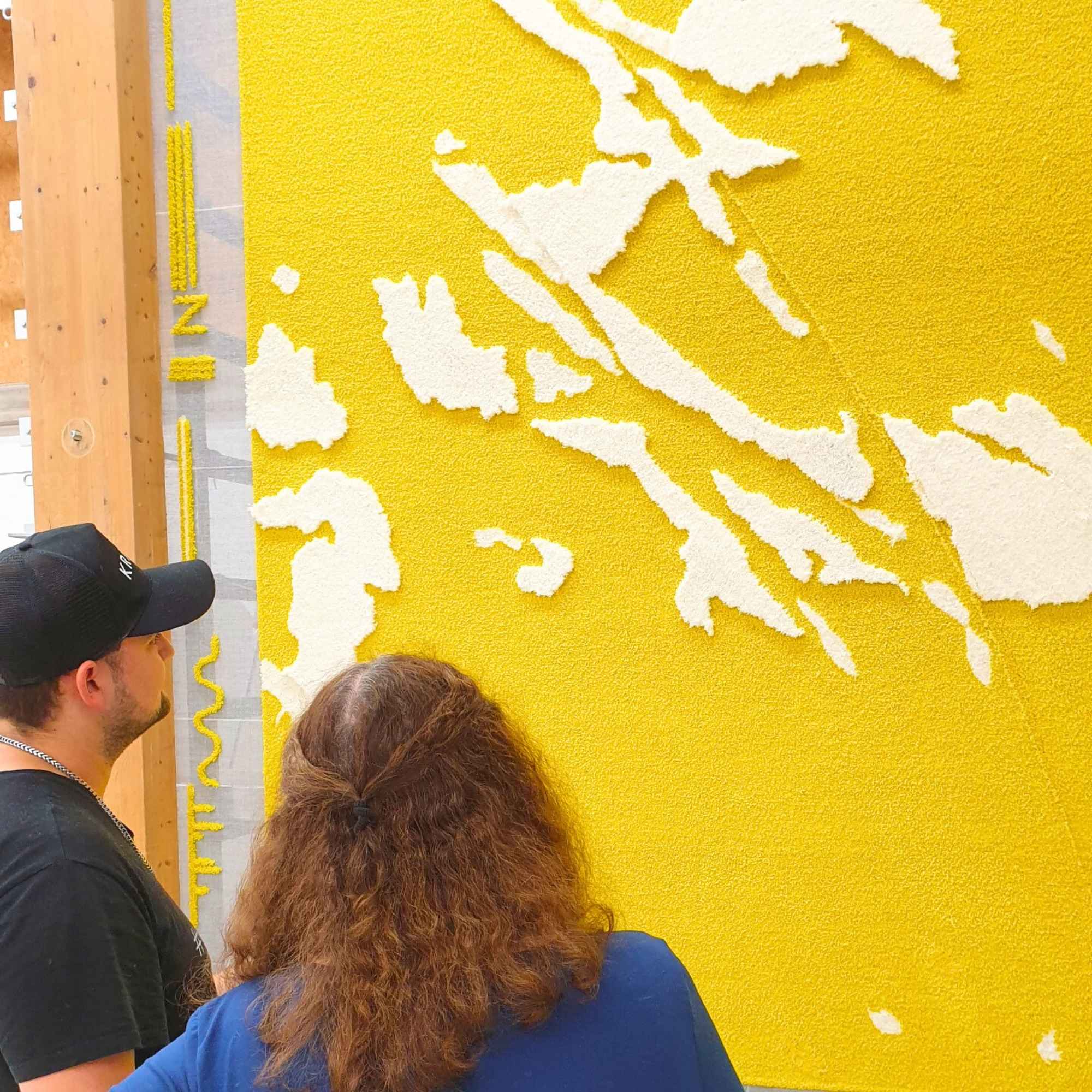 „Zwei Personen betrachten fertigen Wandteppich in Gelb und Weiß an Tuftingwand – Detailphase der Qualitätskontrolle.“
