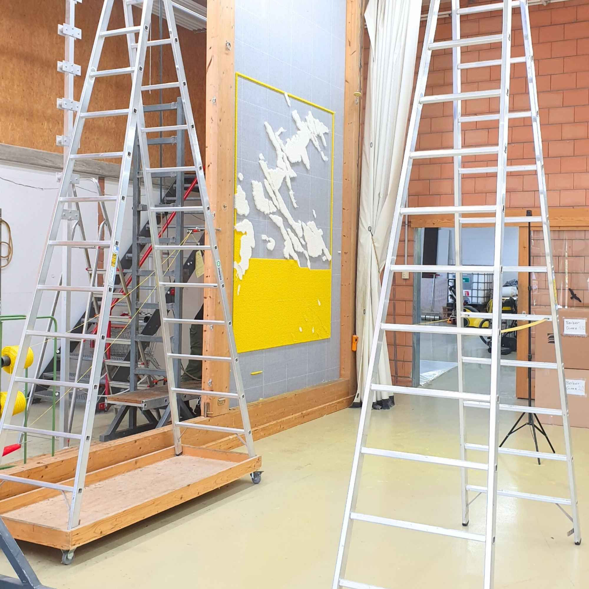 „Teilweise getufteter Wandteppich an senkrechter Arbeitswand, umgeben von Leitern in Werkstattumgebung.“