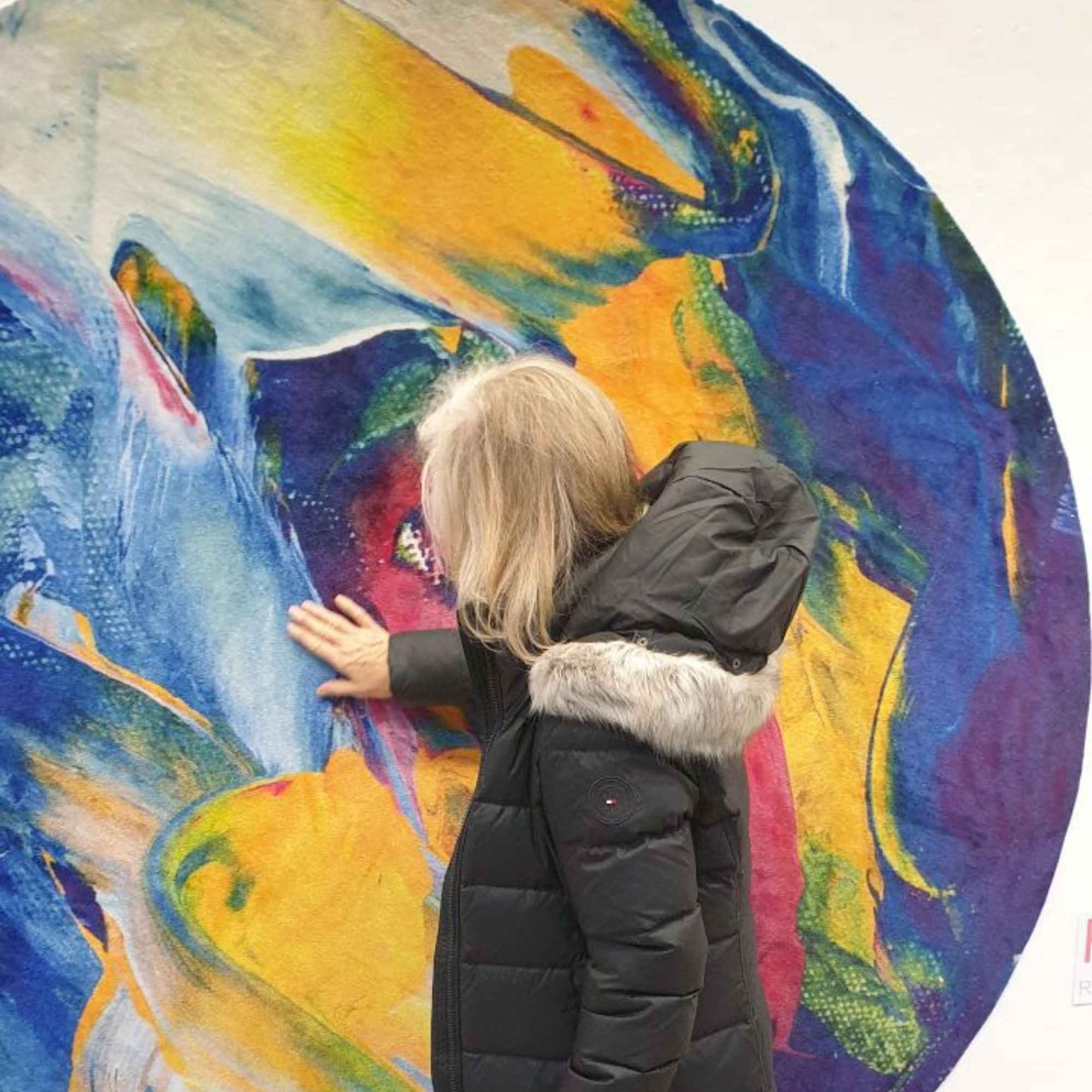 „Besucherin berührt mit der Hand einen runden, farbenfrohen Wandteppich mit abstraktem Motiv.“