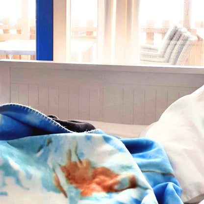 Himmelblaue Kuscheldecke mit weiß-blauen und ockerfarbenen Wolkenmustern liegt auf einem Bett vor einem hellen Fenster.