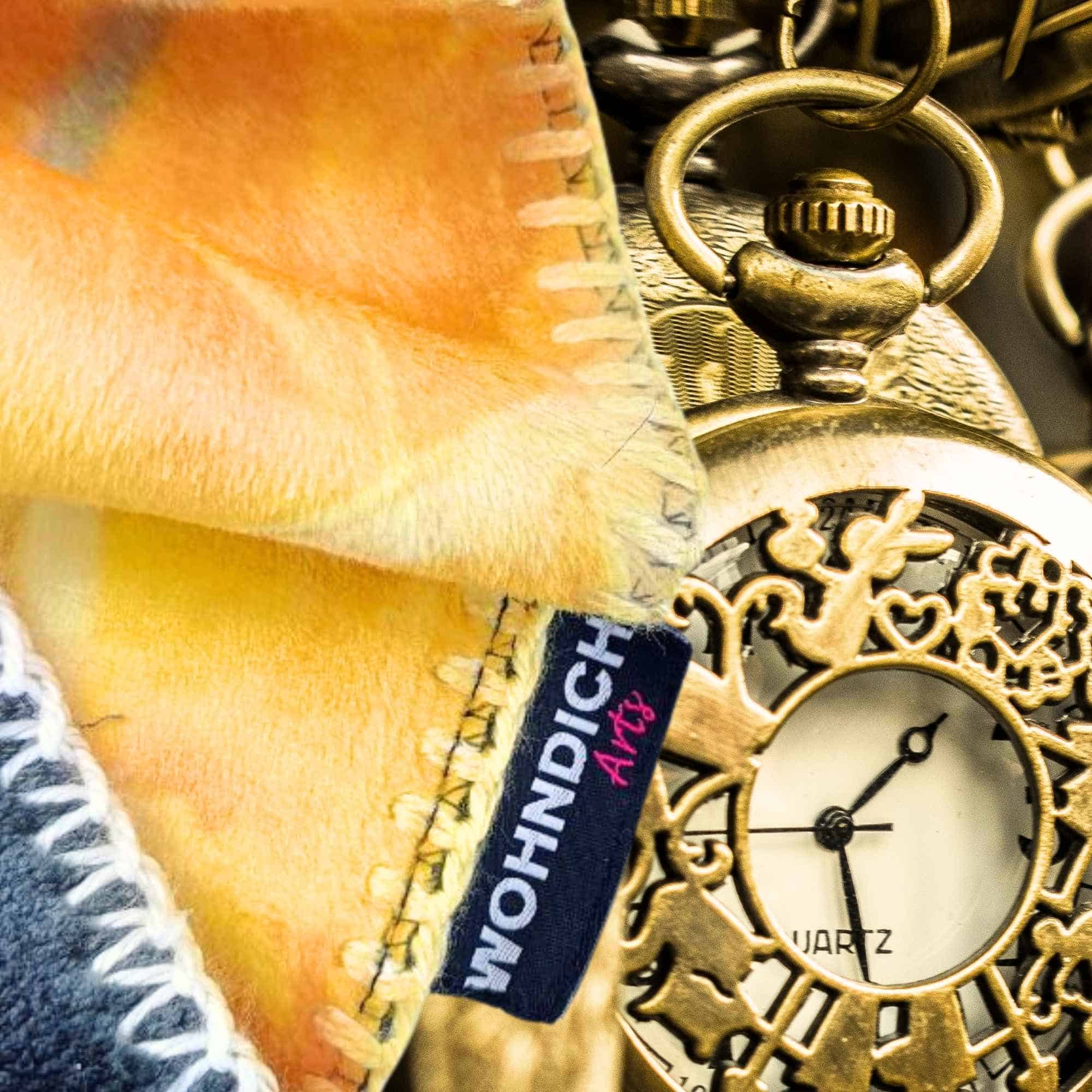 Goldene Taschenuhr neben einer flauschigen Kuscheldecke mit abstraktem Muster in Orange, Gelb und Grau - Phoenix Bundle aus WohnungsReading und Kuscheldecke.