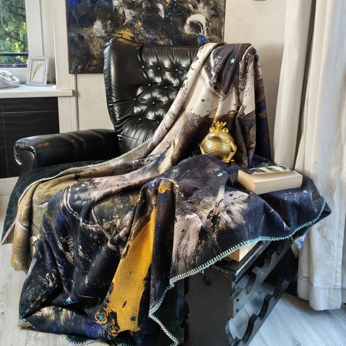 FEEL Kuscheldecke mit abstrakter Kunst in Blau, Schwarz, Weiß und Gold über einem schwarzen Ledersessel, passend zum Originalgemälde von MYSTIQUE - Kunstdecke von INTO THE WOO.