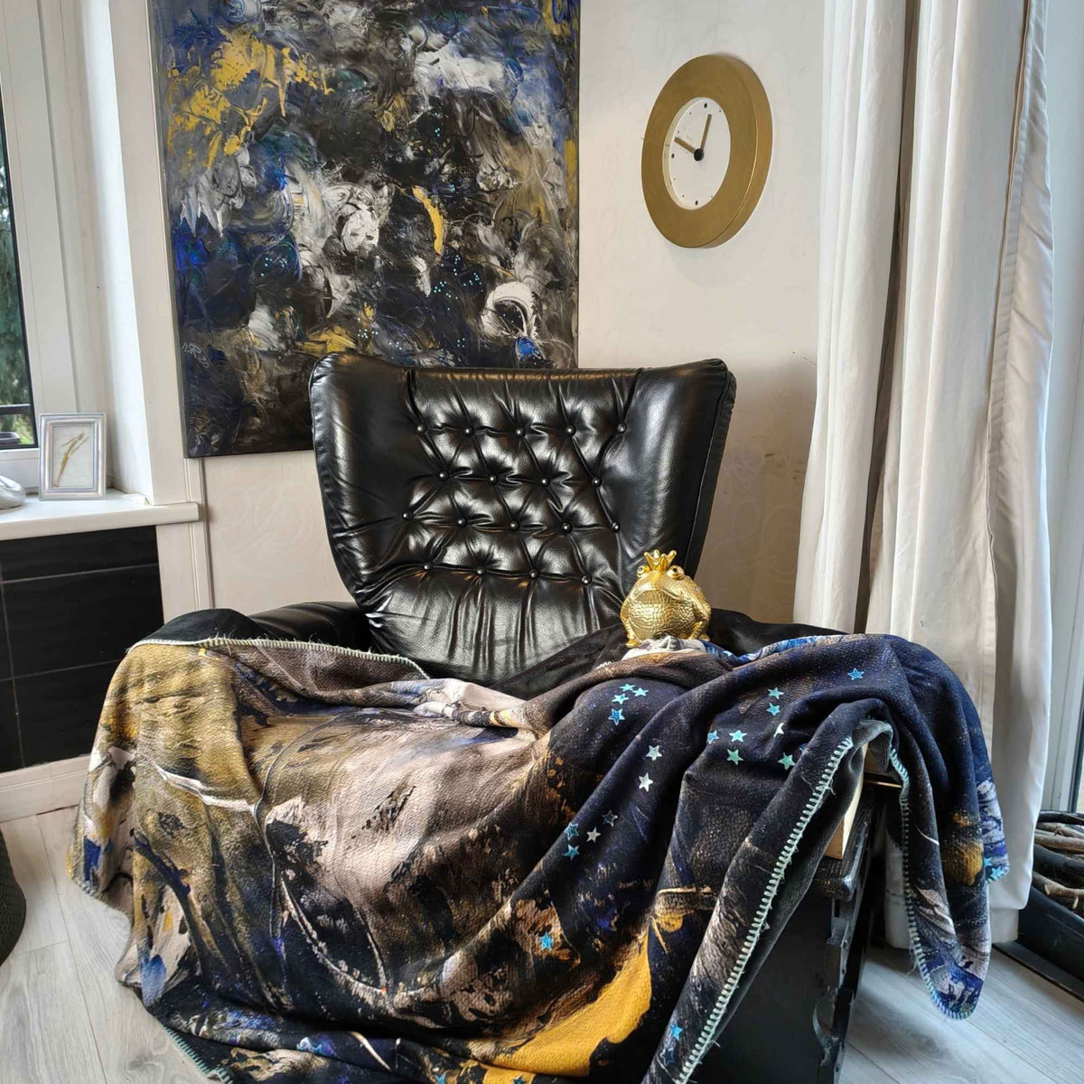 FEEL Kuscheldecke mit abstraktem Kunstmotiv in Blau, Schwarz, Creme und Gold auf einem schwarzen Ledersessel, mit passendem Originalgemälde von MYSTIQUE im Hintergrund - INTO THE WOO.