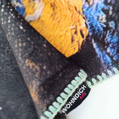 Detailaufnahme einer Kuscheldecke mit sichtbarem „WOHNDICH Arts“-Label, farbigem Kettelrand und strukturreichem Druck in Ocker, Blau und Schwarz.