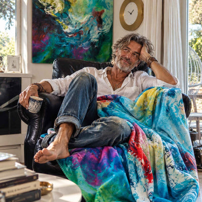 FORREST GUMP Kuscheldecke mit abstrakter Kunst in Türkis, Blau, Grün und Pink: Ein lässiger Mann entspannt barfuß mit einer Tasse Kaffee im Ledersessel. Das farbenfrohe Kunstwerk von MYSTIQUE hängt im Hintergrund.