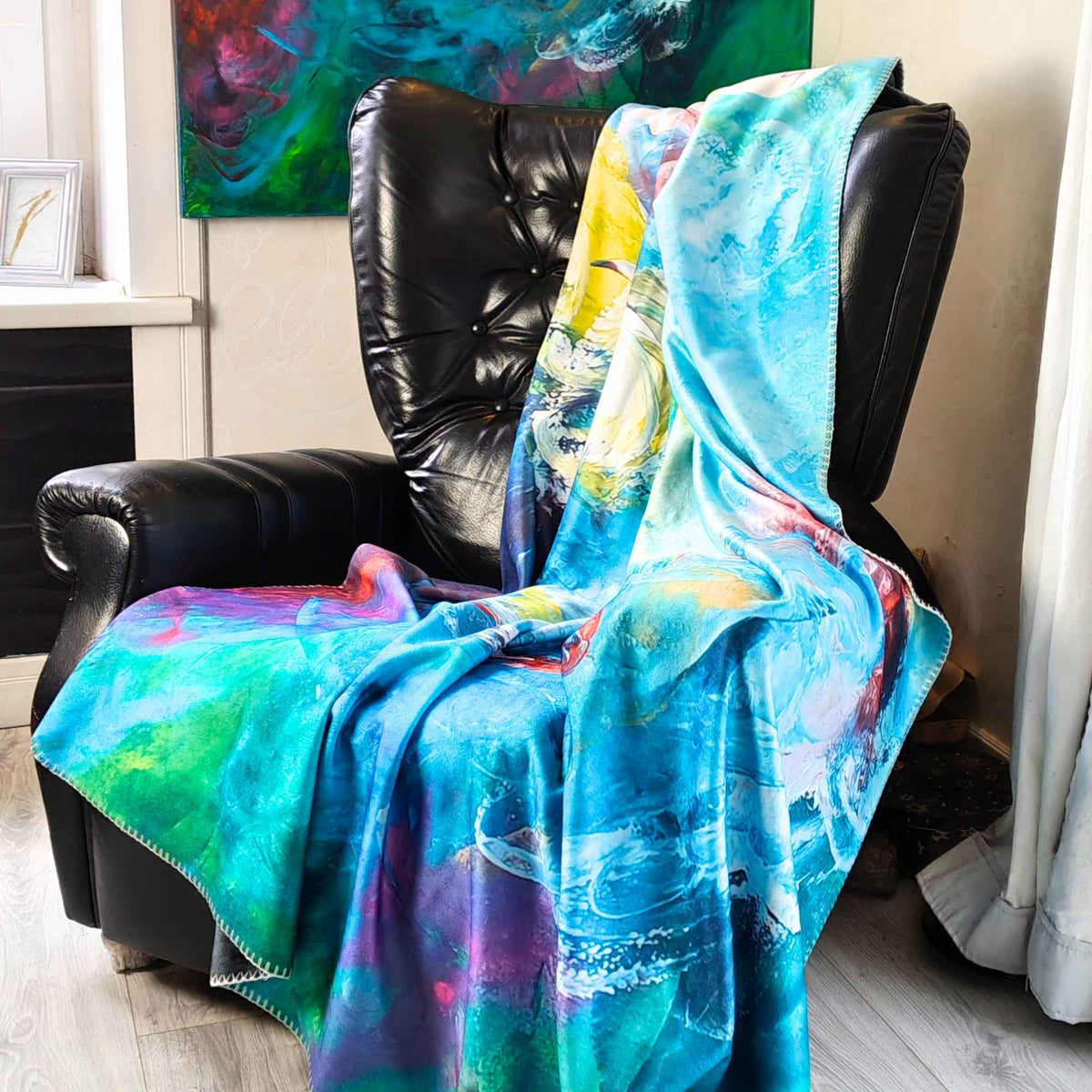 FORREST GUMP Kuscheldecke mit abstrakter Kunst - farbenfrohe Kunstdecke in Türkis, Blau, Grün und Pink auf einem schwarzen Ledersessel. Künstlerische Kuscheldecke nach dem Originalgemälde von MYSTIQUE, erhältlich bei INTO THE WOO.