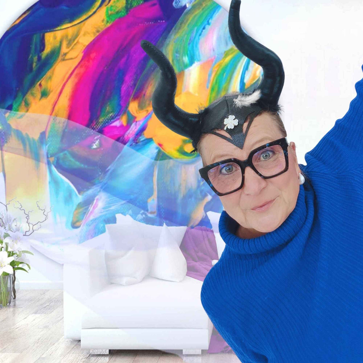 Regine Rauin alias MYSTIC MAVERESS mit schwarzen Hörnern und Brille vor einem farbenfrohen abstrakten Kunstwerk – sie liest Wohnungen und macht mit WohnungsReadings verborgene Muster und blinde Flecken sichtbar.