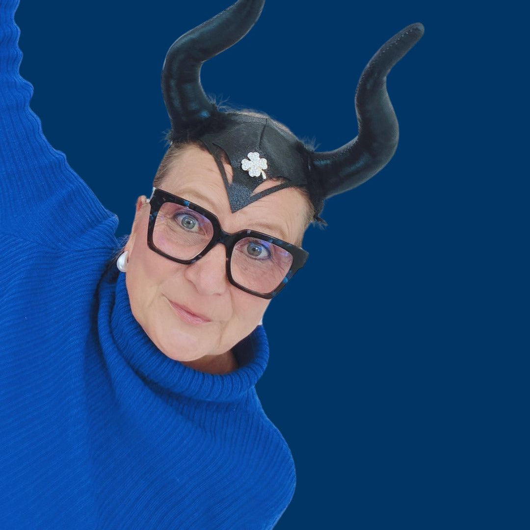 Regine Rauin alias MYSTIC MAVERESS mit schwarzen Hörnern und großer Brille vor dunkelblauem Hintergrund – die Hörner als Symbol für ihre Antennen ins WOO und ihre intuitive Wahrnehmung bei WohnungsReadings.