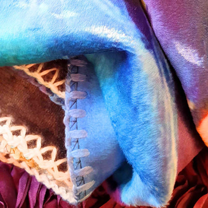 „Detail der farbigen Kuscheldecke mit blauen und violetten Tönen, sichtbare Kettelnaht.“