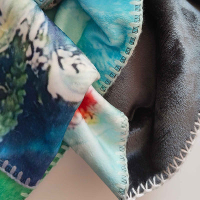 Detail der flauschigen Kuscheldecke FORREST GUMP mit abstraktem Muster in Blau, Türkis, Grün, Weiß und Rot sowie heller und dunkler Stoffseite