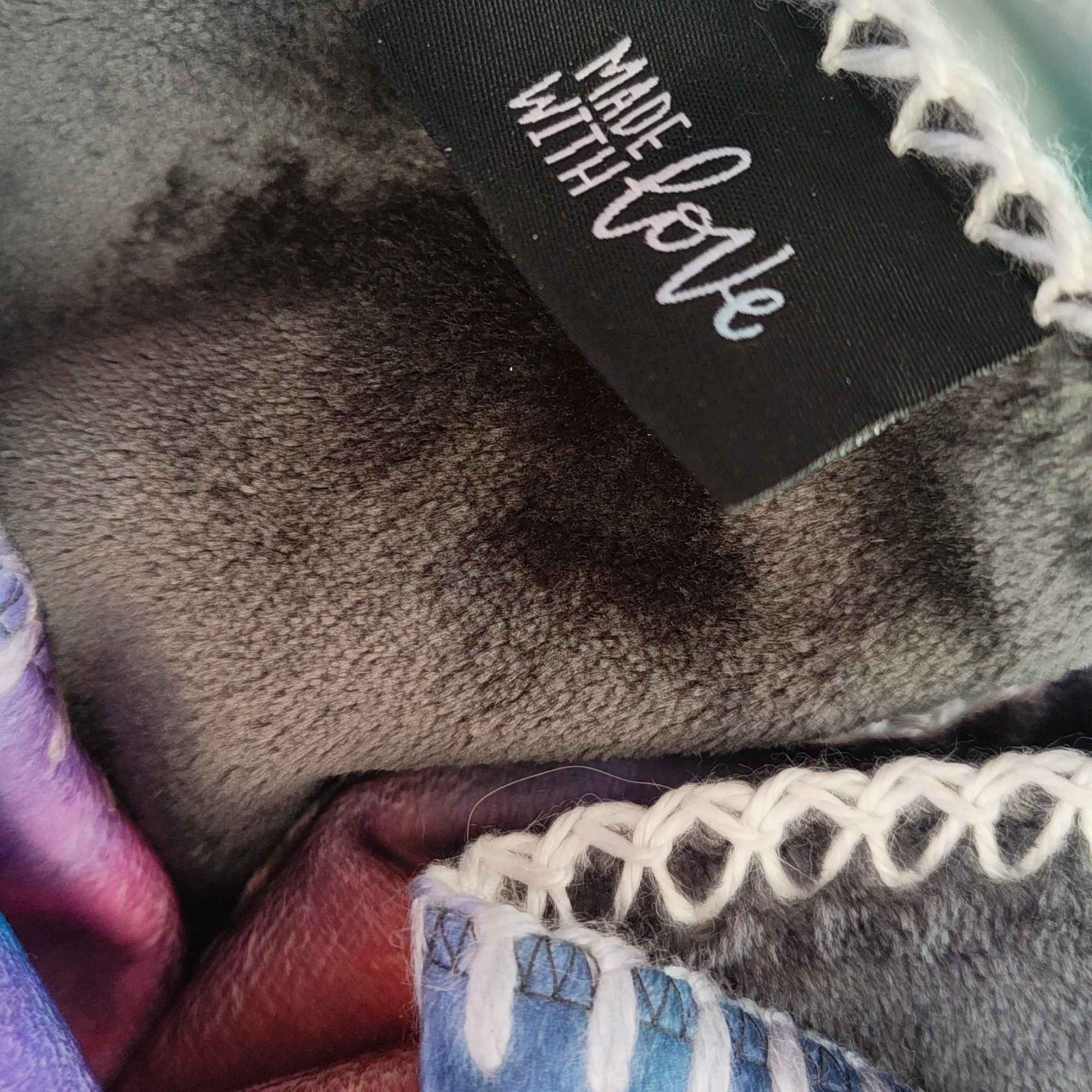 Detail einer weichen Kuscheldecke mit „Made with LOVE“-Label, dekorativer Ziernaht und abstraktem Motiv in Blau, Rot und Violett - FORREST GUMP.