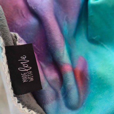 Detail der weichen Kuscheldecke FORREST GUMP mit „Made with LOVE“-Label und abstraktem Motiv in Türkis, Blau, Violett und Rot.