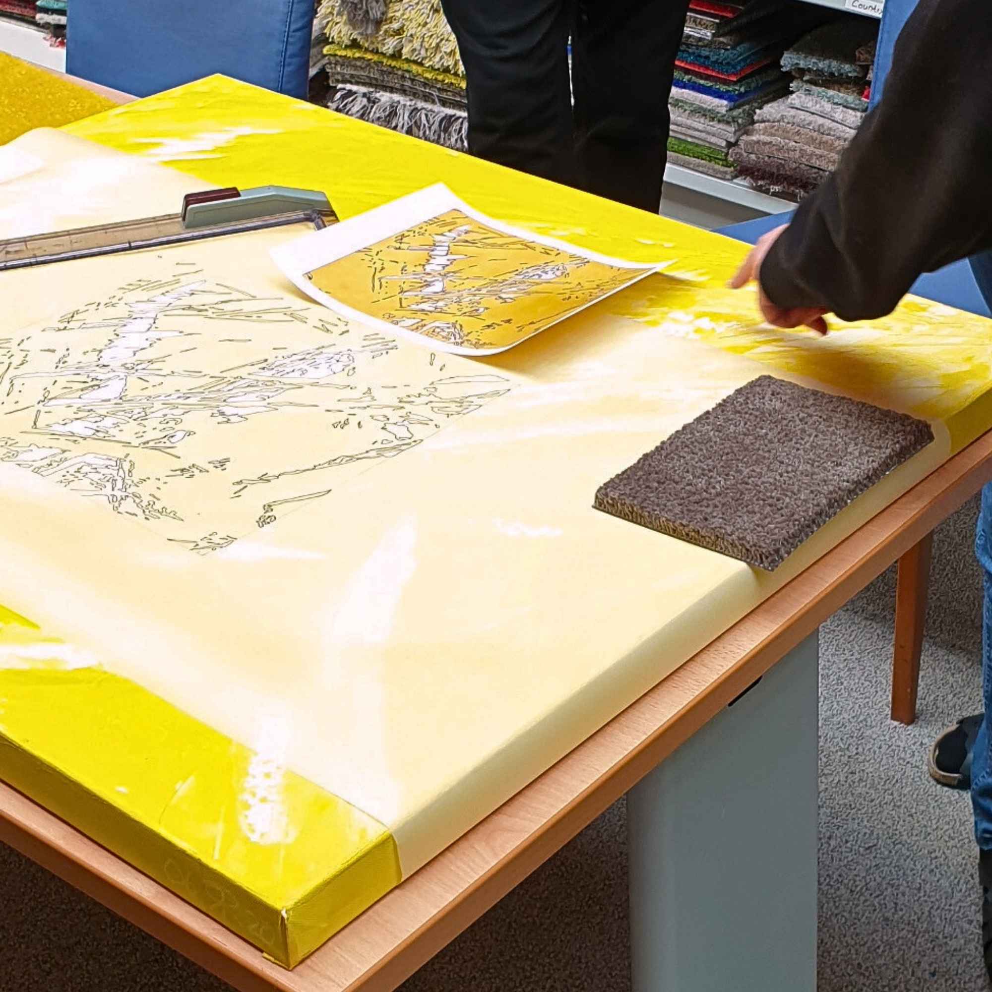 „Entwurf mit Linienzeichnung auf gelbem Bild liegt auf Arbeitstisch, Hände zeigen auf Details, Teppichmusterprobe daneben.“